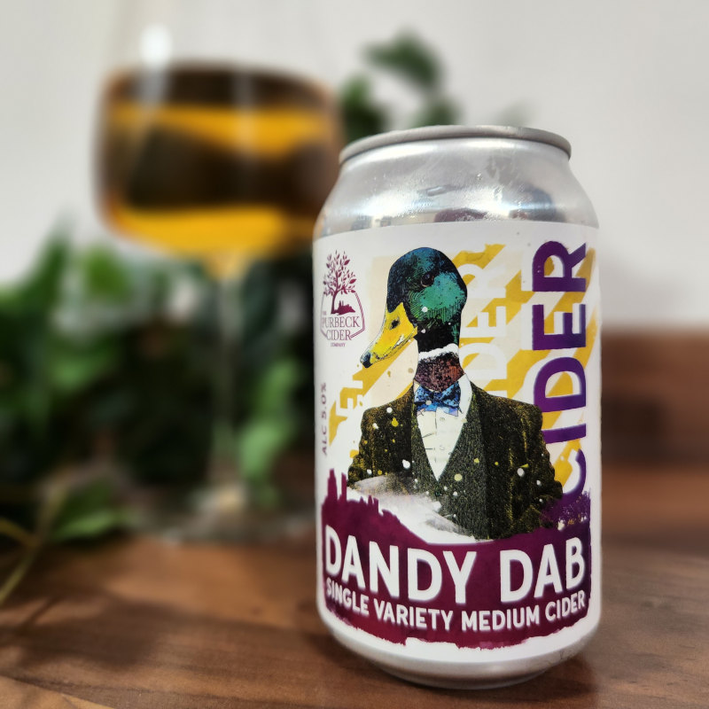 A can of Dandy Dab cider by Purbeck Cider Company. The is a glass with the cider in it blurred in the background.