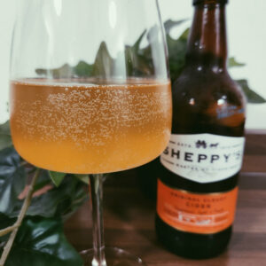 Sheppys original cloudy cider in a wine glass with the bottle in the background.